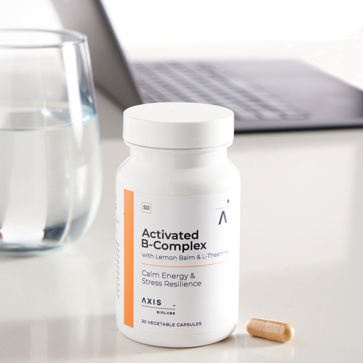 A white bottle of Activated B-Complex dietary supplement with an orange accent stripe and a single capsule lying beside it on a light tabletop, with a blurred laptop and glass of water in the background.